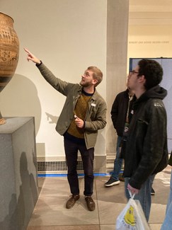 A person with a beard stands in front of some other people and gestures up at a very large ceramic pot on a museum display stand, partly visible at left