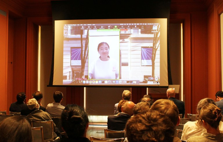 A woman appears on a screen at the front of a lecture hall with a people in the audience