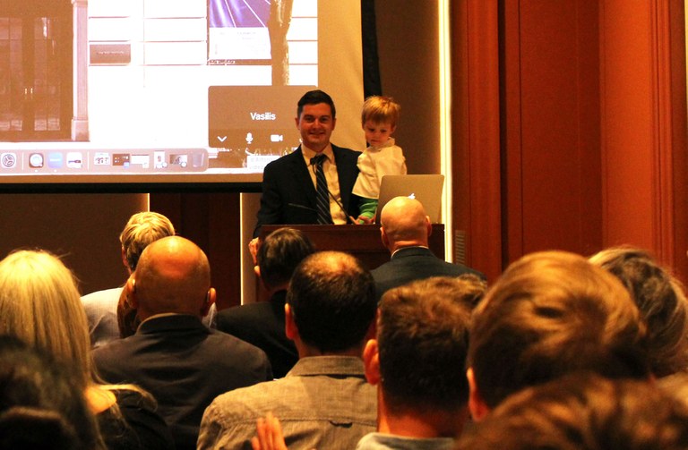 A man and his young son stand at a lectern at the front of a lecture hall with a number of people in the audience.