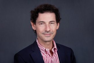 Headshot of Gilles Bransbourg wearing a red-and-white striped shirt and blue blazer against a gray backdrop.