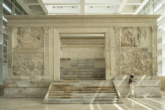 A woman stands in front of a large ancient marble building that is partially restored