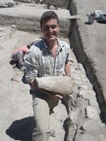 An archaeologist stands amid the excavation, holding a conical stone object.