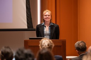 Katherine Thomson stands before a podium and a mic in the ISAW Lecture Hall