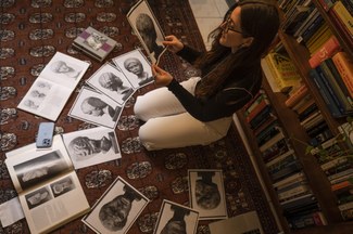 A woman sitting on a red rug on the floor near a bookshelf surrounded by and looking at a series of black and white photographs of an ancient statue.