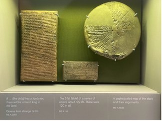 Three artifacts pictured on display, one round, one small and rectangular, and one larger rectangular piece, all with markings on them. Labels at the bottom of the display case read as follows. Left, beneath a tall, rectangular tablet: "If ... (the child) has a lion's ear, there will be a harsh king in the land. Omens from strange births ME K.2007." Center, beneath a smaller rectangular tablet: "The 61st tablet of a series of omens about city life. There were 120 in all. ME K.116." Right, beneath a large circular tablet: "A sophisticated map of the stars and their alignments. ME K.8538."