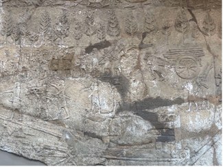 A close up image of an ancient wall full of relief markings.  Legible shapes include: trees, human figures in large groups (some holding weapons or tools), a cart full of logs pulled by human figures, and a boat carrying what appears to be a large sculpture of a bull or other creature, laid on its side.