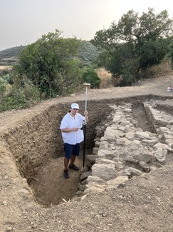 A man in a white shirt and white cap stands in a deep, polygonal excavation with very straight sides. Next to him, a massive stone structure, possibly a fortification wall or building platform, can be seen. He is holding a tall pole with a flat disk on its top. Behind him, some trees are visible, as well as a tree-covered hill in the distance.