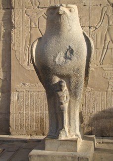 Photograph of a stone statue of a large falcon with a small human figure standing between its legs. In the background is a wall covered in hieroglyphic inscriptions.
