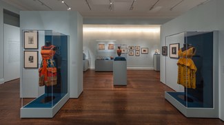 A gallery room with artifacts on the blue walls and in glass cases including an ancient vase and two colorful costumes front and center.