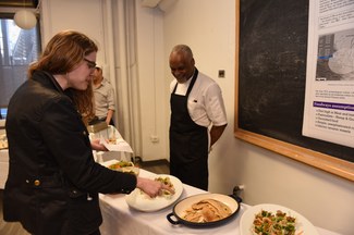 A chef, wearing an apron, stands behind a table bearing several different dishes of food while other people examine the dishes.