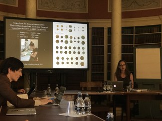 Scholars with laptops in a conference room in front of a large projection screen displaying a picture of ancient coins.