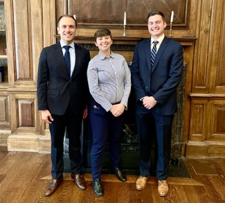 Three individuals pose in front of a fireplace in a formal library