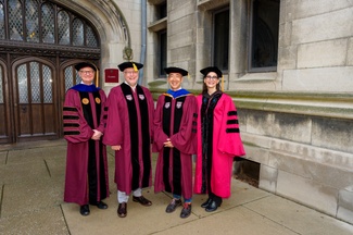 Four faculty members wearing maroon and pink academic robes and caps pose outside a Gothic-style stone building during a convocation ceremony.