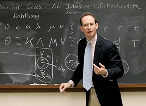 A man in a jacket and tie stands in front of a chalkboard, apparently addressing a class. On the chalkboard is written "Ancient Greek An Intensive Introduction" and below that text appears the word "dipthong" and a listing out of the Greek alphabet. 