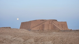 The Great Ziggurat of Ur, a massive Mesopotamian structure, dedicated to the Moon God is pictured against a pale blue dusk sky with a full moon behind it. 
