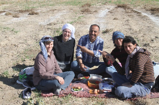 Five people sitting on a blanket in a field eating from portable containers.