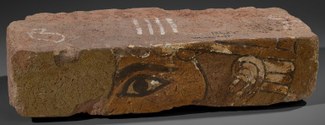 A rectangular brick with a part of a god's face on the face of the brick. The eye and ear are prominent. The brick is light brown with hints of black, white, and light green pigment