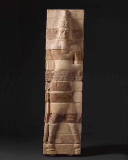 Rectangular pillar with bricks in molded relief showing a man with a long beard and a tall hat, as well as bull-like hooves for legs. The whole figure is divided into 14 stacked horizontal bricks and the brick at the mid section has a cuneiform inscriptio