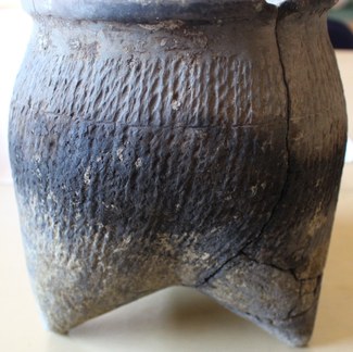 Photograph of a round ceramic vessel with a broad, open rim, stippled texture, and three legs that emerge seamlessly from the body of the vessel and terminate in narrow points. 