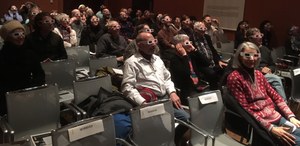 A group of people in a darkened lecture hall wear 3D glasses.