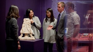 A group of people stand around a pyramidal stone object on a display stand. One gestures at the object. Another display case containing gold jewelry can be seen behind them.