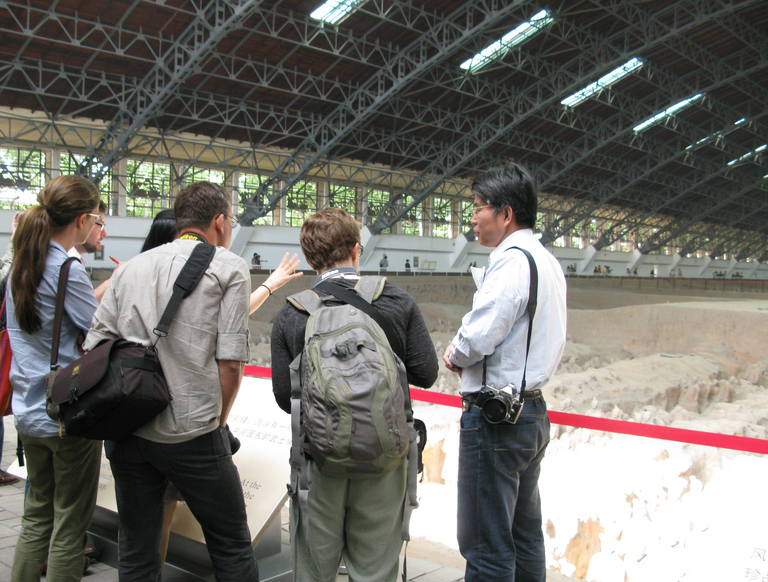 A group of people with backpacks and cameras are photographed from behind. They stand at a glass retaining wall in front of an excavated pit of ceramic warrior statues. The arched protective roof over the excavated area is also clearly visible. Photo courtesy of Lillian Tseng.