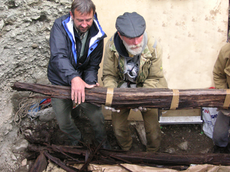 Two men grasp and lift a large wooden beam that extends out of the photo to the right. They appear to be in an excavated trench. Both are wearing warm outer garments.