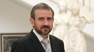 Photograph of a man wearing a suit and jacket. In the background, a part of a framed painting and of a classicizing sculpture of two female figures can be seen.