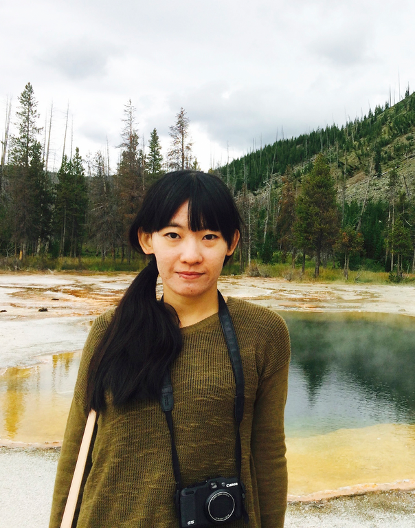 A woman with a camera on a strap around her neck looks into the camera. Behind her can be seen portions of a landscape with a body of water, sandy shore area, coniferous trees, and part of a hillside covered in vegetation.