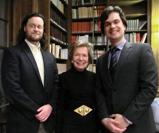 Three people in semi-formal attire stand in a wood-panelled room with bookshelves and smile for the camera.