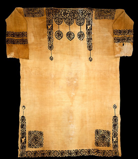 A short-sleeved tunic made of yellowish material is photographed lying flat. Elaborate patterns in a dark color adorn the top and bottom of the garment, while also ringing the sleeves.