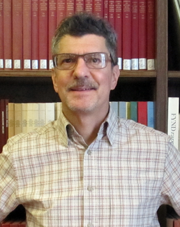 A man wearing glasses smiles for the camera while standing in front of a book shelf.