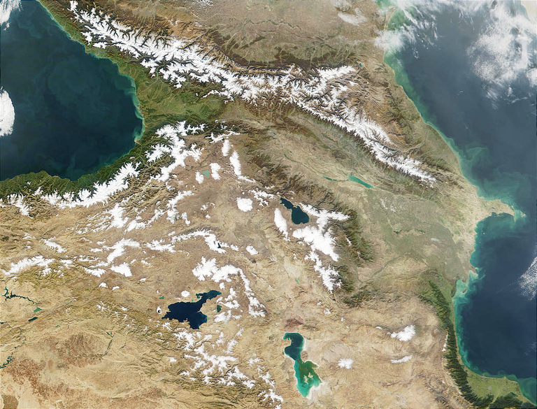 A satellite photo centers on a mountainous region with snow-capped peaks running from northeast to southwest, bordered on both west and east by parts of bodies of water.