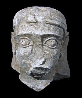 A photograph of a sculpted gray stone head on a black background. The nose and other parts have broken off.