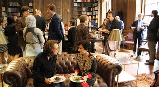 Several groups of people in semi-formal attire carry plates of food while sitting or standing in a wood-paneled room with bookshelves. Bright daylight floods the room from two large windows on the right.