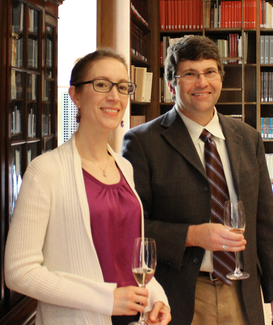 Two people in semi-formal attire stand and smile for the camera in a wood-panelled room with bookshelves. 