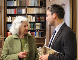 Two people stand talking and hoding wine glasses in a wood-panelled room with bookshelves.