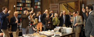 A large group of people in semi-formal attire stand in a wood-paneled room with bookshelves. They surround a rectangular table laden with serving trays of food. They raise wine glasses in a toasting gesture.