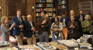 A large group of people stand in a wood-paneled room with bookshelves along and behind a table filled with food. A woman on the left raises a wine glass in a toasting gesture.