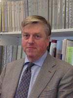 A man wearing a suit and tie looks into the camera. There is a wall of bookshelves behind him.