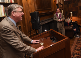 A man in a suit and tie speaks from a podium. The photograph is taken from the side and a bit behind him. Beyond him, one can see a wood-paneled room with bookshelves and a large fireplace. Another man stands, and a woman sits, listening.