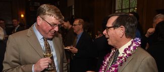 Two men wearing suits and holding wine glasses smile and speak to each other. The man on the left holds a glass of white wine. The man on the right is wearing a necklace of purple and white flowers.