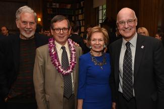 Four people stand next to eachother smiling for the camera. One of the men wears a necklace of white and purple flowers. They are in a wood-paneled room with bookshelves and windows.