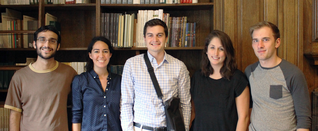 Five people wearing informal dress pose standing for a photograph in a wood-paneled room with bookshelves behind them.