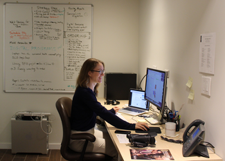 A woman sits at a desk on which several computers, monitors and other technical devices can be seen. On the wall behind, a whiteboard holds notes in multiple colors of ink. On the floor below it, another computer sits with keyboard and other components stacked on top of it.