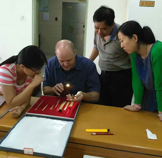 A large shallow box lined with red padding stands open on a table and holds several long, narrow objects that are light beige in color. A man sits in front of the box and holds an object in his hand. He aims a light at the object with his other hand. A seated woman on the left and two standing people on the right look closely at the object in his hand.