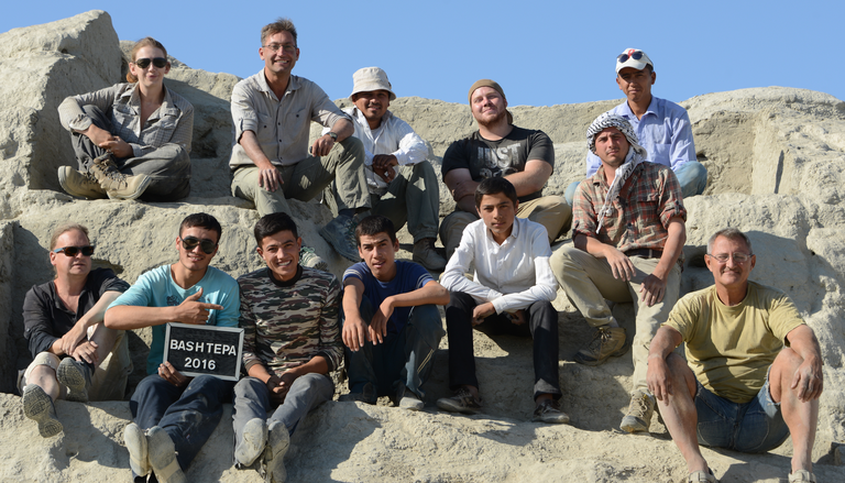 A group of 12 people site on a sloping archaeological site with obvious cuts in the earth. One of them holds a sign reading "Bashtepa 2016".