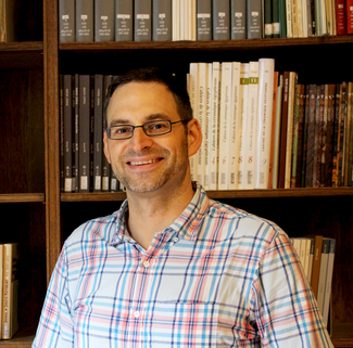 A man smiles into the camera in front of a wall of bookshelves.