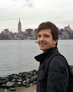 A young man stands, smiling, on a rocky shoreline. The Manhattan skyline--with the Empire State Building prominent--is visible in the distance.