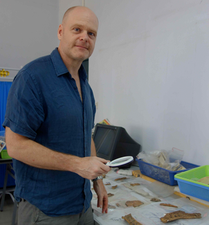 A man stands and looks at the camera. He holds a magnifying glass in one hand. To his side there is a large table covered with artifacts, tools, and boxes.